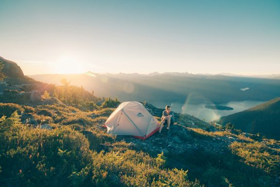 A tent set up on a hillside at sunrise, with a person nearby overlooking a scenic view.